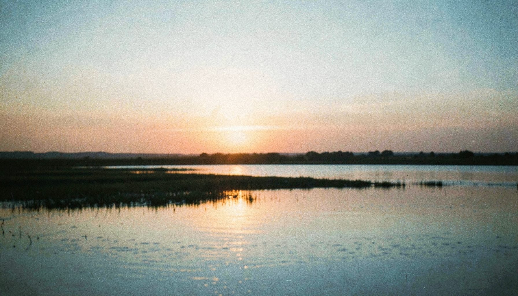 Un ciel de Camargue au coucher du soleil, teinté d'orange doux et de bleu pâle, avec une étendue d'eau calme reflétant la lumière dorée. Une ambiance sereine et commémorative.