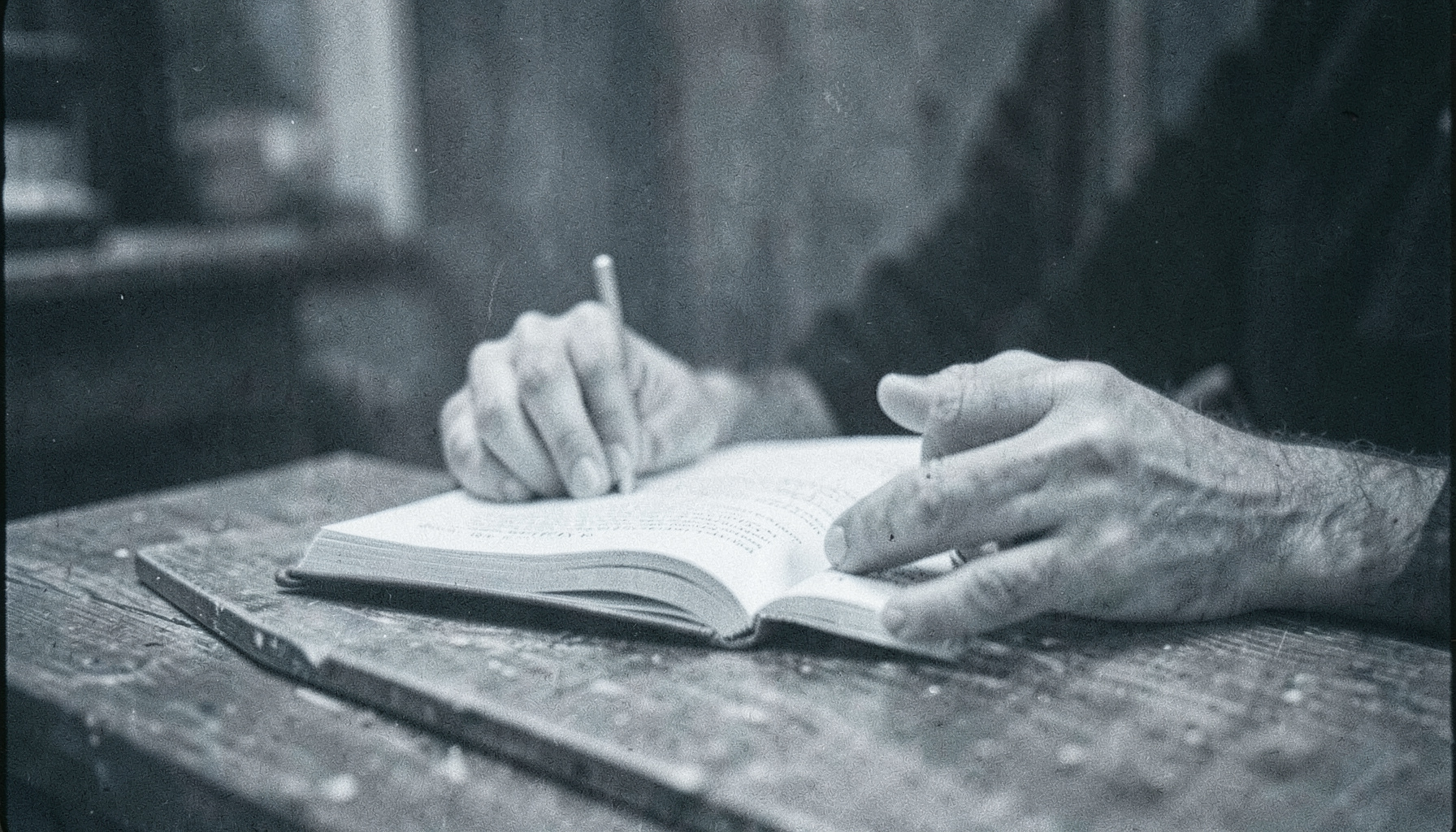 Photographie argentique en noir et blanc granuleux, mains feuilletant un livre ouvert sur une table en bois, lumière douce et contrastée, ambiance studieuse et bienveillante.