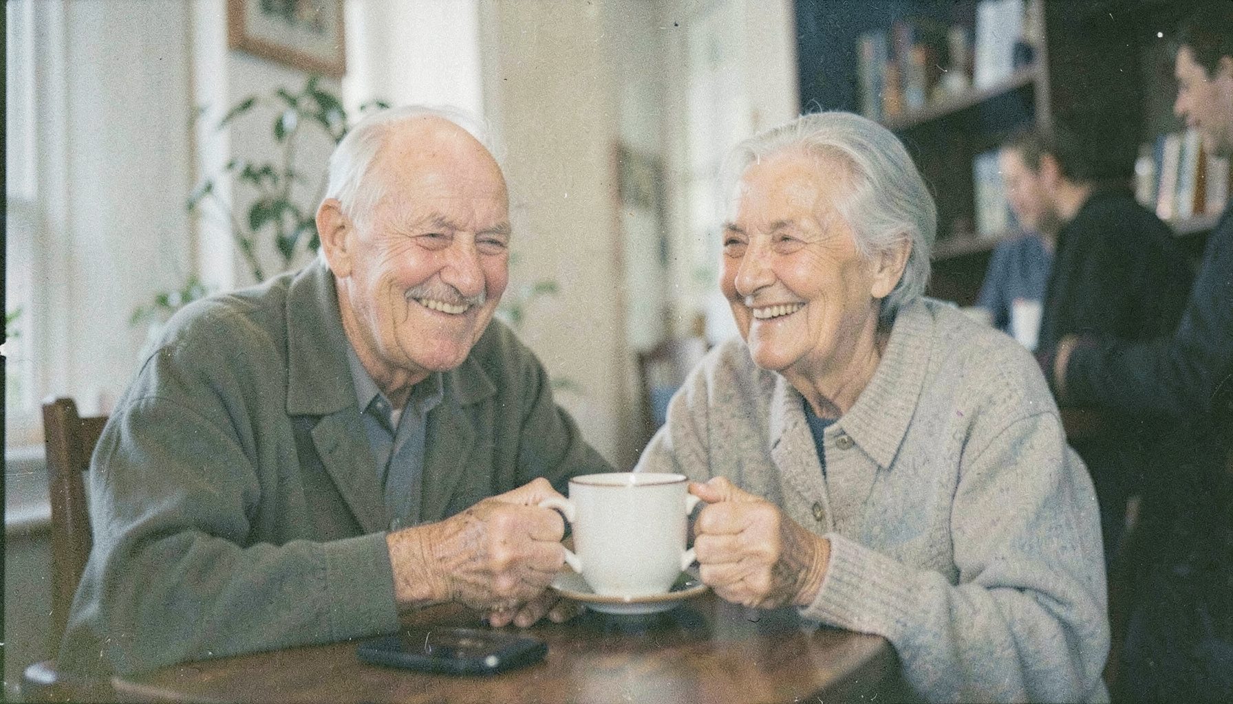 Photographie argentique aux teintes douces et chaudes, grain visible, montrant deux personnes âgées souriant autour d'une tasse dans un cadre convivial, lumière naturelle tamisée.
