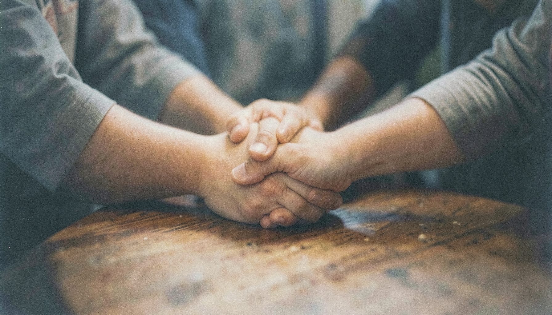 Photographie argentique aux tons chauds et légèrement granuleuse montrant des mains jointes en cercle autour d'une table en bois, évoquant la solidarité et l'engagement collectif dans une ambiance douce et intime.
