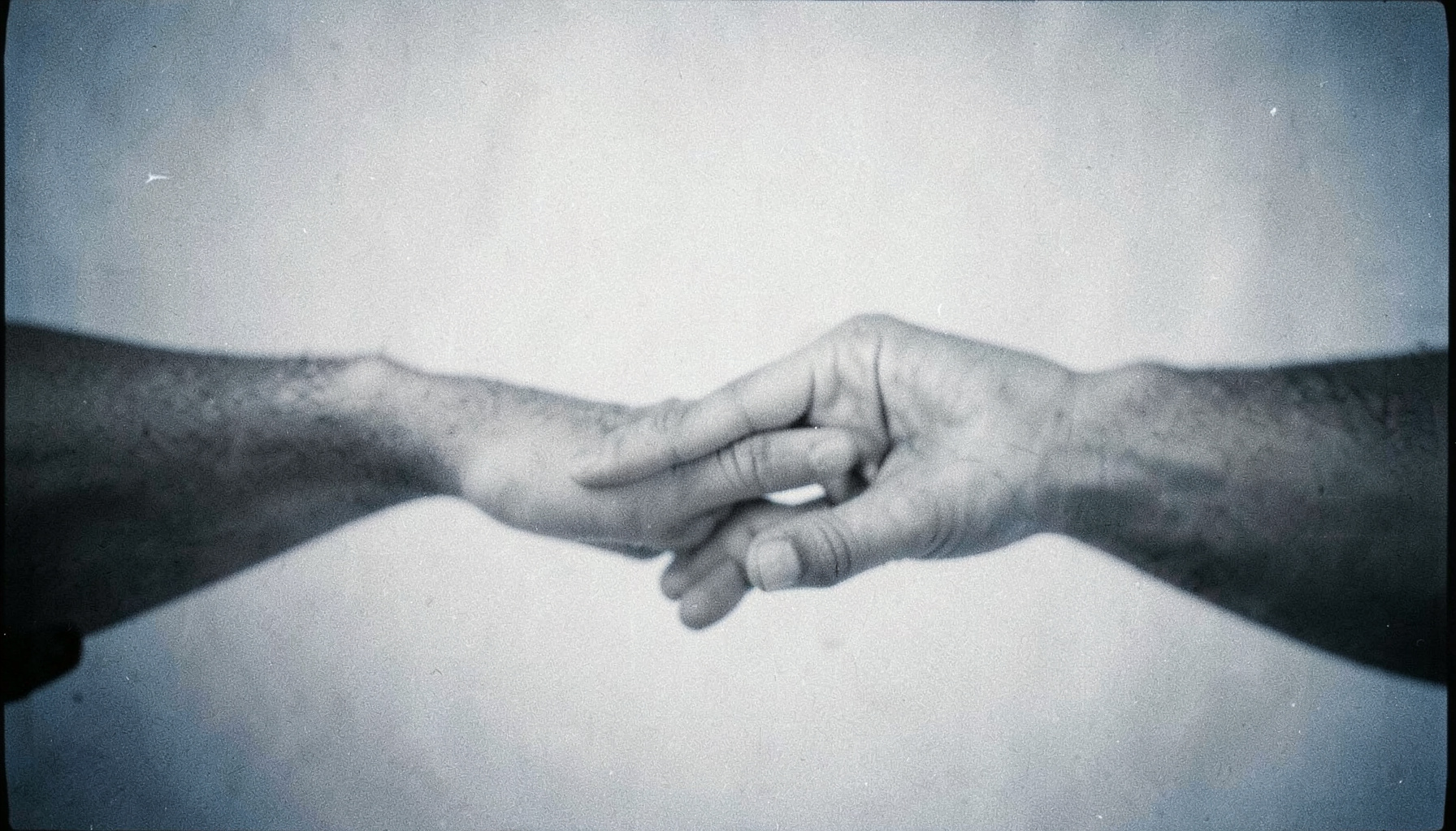 Photographie argentique en noir et blanc d'une main tendue vers une autre, symbole de solidarité et de don, sur un fond doux aux tons gris argenté avec un grain visible.